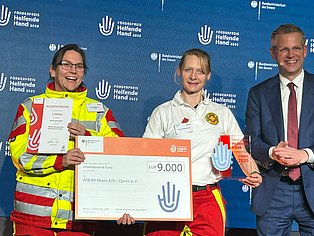 Zwei Personen in ASB-Kleidung halten einen Check, den Preis und eine Urkunde in den Händen. Eine dritte Person im Anzug steht daneben.