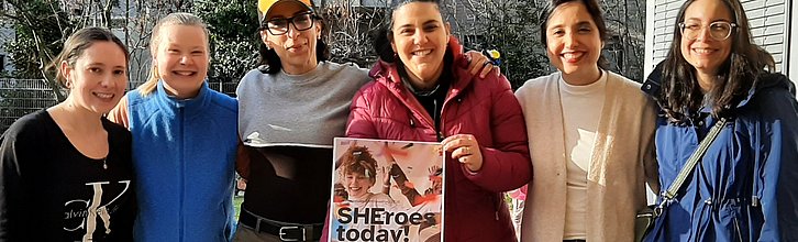 Sechs Frauen verschiedener Herkunft, die glücklich in die Kamera gucken. Eine hält das Plakat "SHEroes today!" in den Händen.