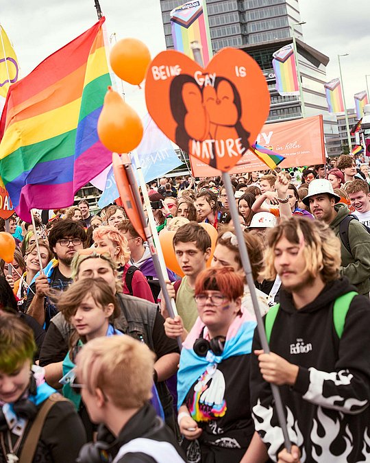 Menschenmenge bei der CSD-Parade. Im Vordergrund ein Schild mit küssenden Pinguinen. Aufschrift: "Being gay is nature"