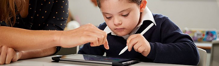 Eine Integrationshelferin sitzt neben einem Jungen in der Schule und zeigt ihm was auf dem Tablet