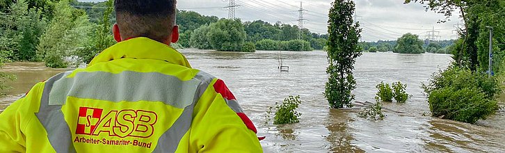 Person in ASB-Arbeitsjacke steht vor einem überfluteten Fluss.
