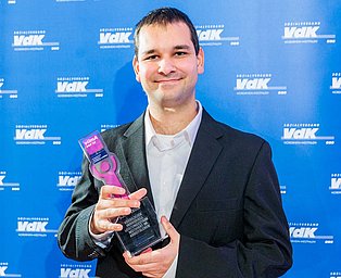 Foto des Preisträgers Björn Esser. Björn Esser lächelt vor dem blauen VdK-Hintergrund und hält den Preis in beiden Händen.