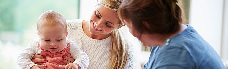 Eine erwachsene Person mit Kleinkind auf dem Schoß. Das Kleinkind spielt mit der Hand einer anderen erwachsenen Person, die daneben sitzt.