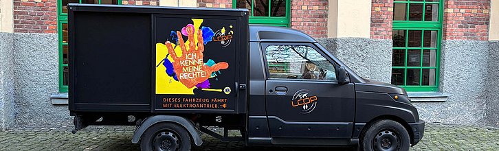 Ein schwarzer kleiner Kastenwagen vor einer Häuserwand. Auf dem Wagen ist eine bunte Grafik mit einer orangenen Hand in der Mitte.