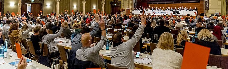 Abstimmung in einem Saal. Personen heben rote Karten in die Höhe.