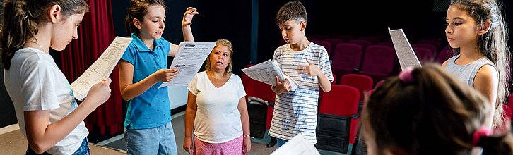 Kinder stehen auf einer Bühne und üben ein Theaterstück. Eine erwachsene Person steht dabei und sieht zu.