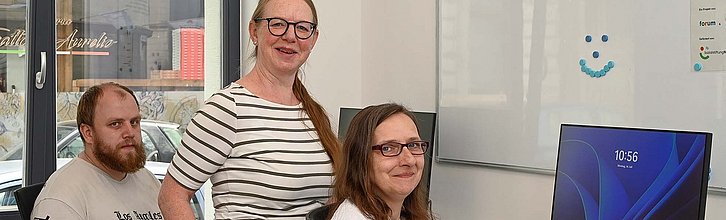 Drei Personen gucken in die Kamera während sie an Arbeitsplätzen mit Desktop sitzen/stehen.