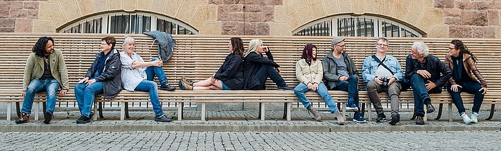 Eine bunte Gruppe aus Menschen sitzt auf einer großen Holzbank in verschiedenen Positionen.