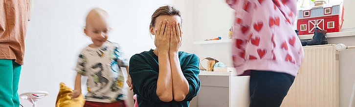 Eine erwachsene Person schlägt die Hände vors Gesicht. Um sie herum toben Kinder in einem Spielzimmer.