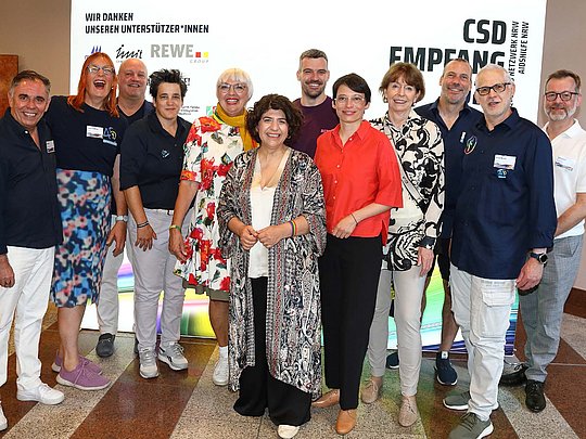 12 Personen vor einer Werbetafel des CSD-Empfangs. U.a. Claudia Roth und Henriette Reker sind zu sehen.