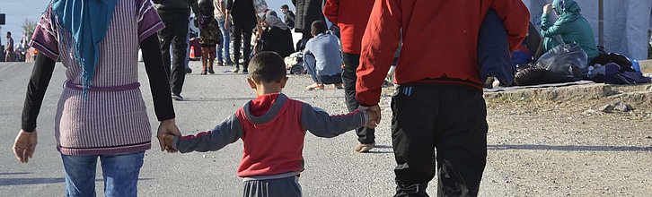 Zwei Erwachsene halten in ihrer Mitte ein Kind an den Händen und laufen durch ein Flüchtlingslager