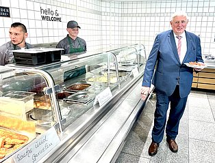 Minister Laumann steht in einer Kantine. Hinter der Essensausgabe stehen zwei Projektteilnehmende.