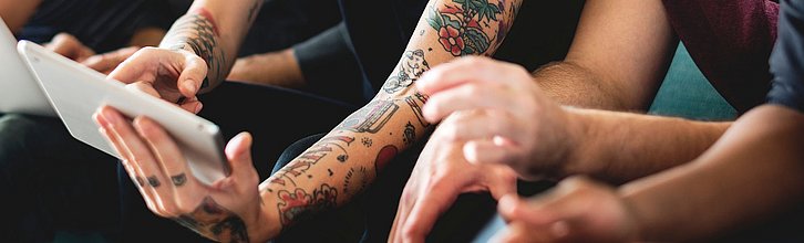 Drei Oberkörper von Erwachsenen, die nebeneinander sitzen. Person in der Mitte hält ein Tablet in den Händen.