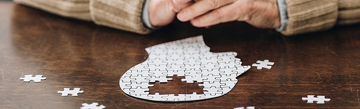 Ein Kopf aus Puzzleteilen. In der Mitte fehlen Teile. Im Hintergrund zwei faltige Hände.