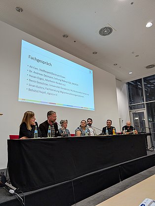 Sieben Personen sitzen auf einer Bühne an einem Pressetisch und unterhalten sich.