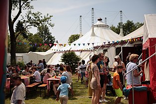 Menschen in sommerlicher Kleidung sitzen und stehen vor dem Zirkuszelt. Das Gelände ist mit Wimpelgirlanden geschmückt.