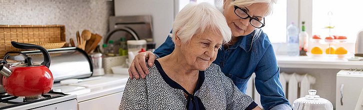 Eine alte Person sitz am Küchentisch während eine jüngere Person unterstützend hinter ihr steht und ihr eine Hand auf die Schulter legt.