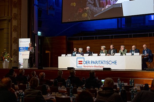 Blick auf das Podium mit Aufsichtsrat und Vorstand des Paritätischen NRW, Elke Schmidt-Sawatzki am Rednerpult