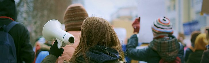 Demonstrationszug von hinten. Eine Person hält ein Megafon in der Hand. Andere halten Plakate.