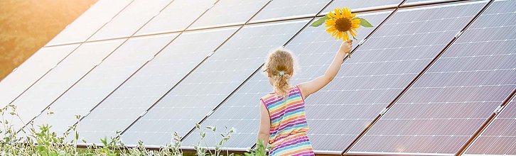 Ein Mädchen mit einer Sonnenblume läuft auf einem Feld entlang von großen Solarpanelen.
