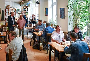 Ein volles, einfach möbliertes Restaurant. An allen Tischen sitzen Menschen die Essen. In der Mitte stehen zwei sich unterhaltende Personen