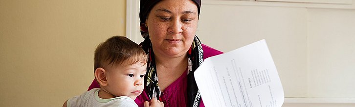 Eine Frau mit Kopftuch hält ein Kleinkind auf dem Arm und schaut Papiere durch.