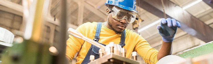 Eine junge erwachsene Person steht mit Helm und Schutzbrille vor einer Maschine