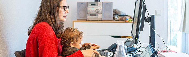 Eine Mutter sitzt mit ihrem Kind auf dem Schoß am Schreibtisch vor einem Computer