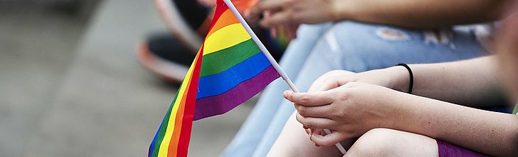 Jugendliche sitzen auf einer Treppe. Eine Person hält eine Pride-Flagge in den Händen.