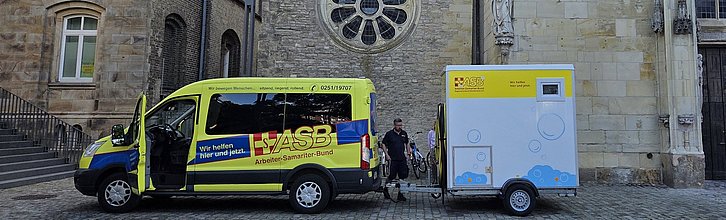 Transporter mit ASB-Logo samt Duschanhänger vor einem Kirchengebäude.
