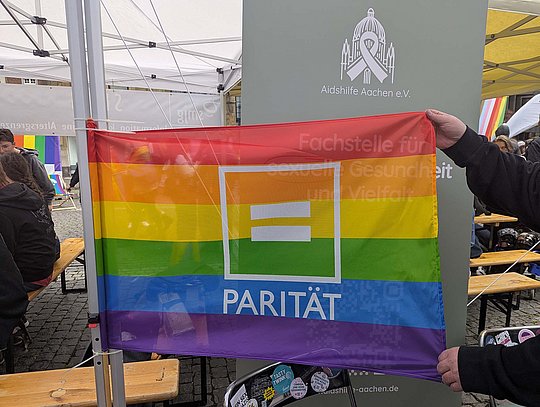 Parität-Flagge in Regenbogenfarben. Im Hintergrund ein Roll-Up der Aidshilfe Aachen.