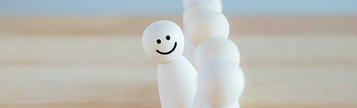 Eine Holzfigur mit lächelndem Gesicht blickt aus einer Reihe mit anderen Holzfiguren hervor.