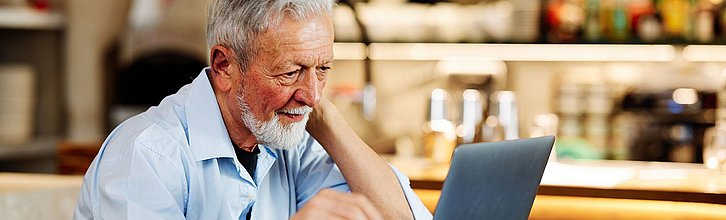 Ein älterer Mann sitzt mit einer Tasse Kaffee vor seinem Notebook und schaut interessiert  auf den Bildschirm.