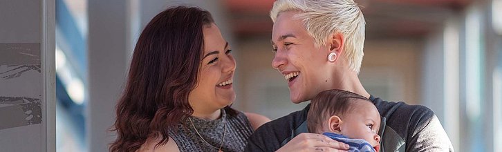 Zwei erwachsenen Personen, eine langhaarig die andere kurzhaarig, lächeln sich an und halten ein Baby auf dem Arm.